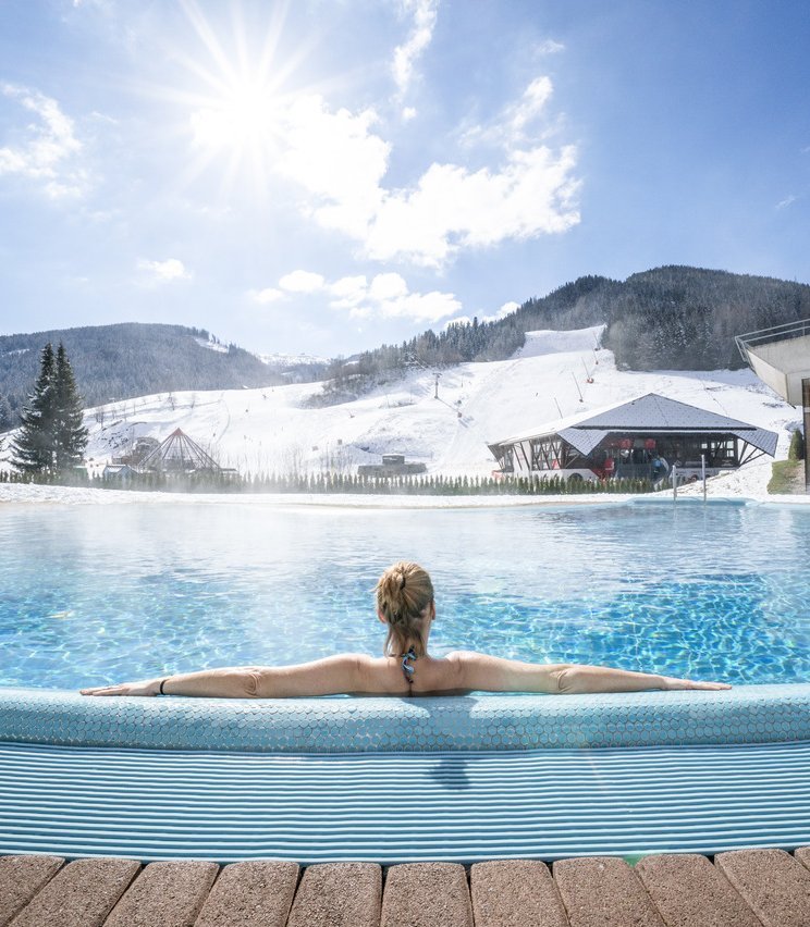 Winterlicher Poolgenuss Person entspannt im beheizten Außenpool mit Blick auf verschneite Berge bei Sonnenschein.